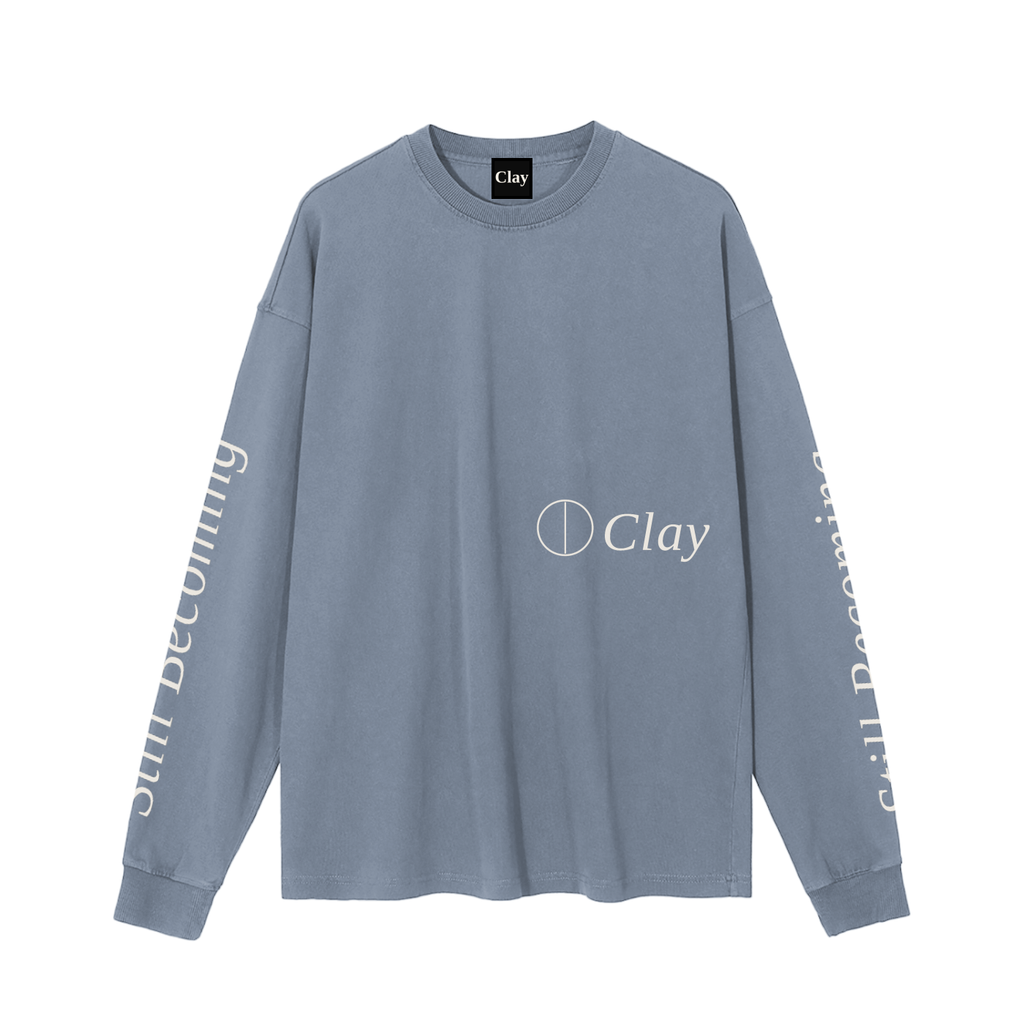 The Gatlinburg Long Sleeve T-Shirt is a unisex cotton top in faded blue, featuring “Clay” and a circle logo on the chest plus “Still Becoming” down both sleeves—a casual streetwear choice for autumn and winter.