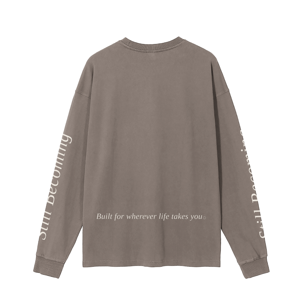The Gatlinburg Long Sleeve T-Shirt is a street-casual, taupe top with “Built for wherever life takes you” on the lower back and “Still Becoming” in light serif lettering down both sleeves.